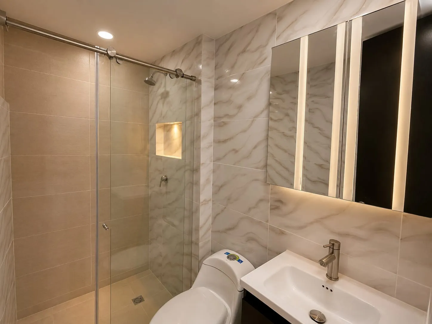 Baño de inspiración moderna, donde los acabados tipo mármol, la iluminación integrada y la división en vidrio crean una atmósfera elegante, funcional y sofisticada. Un espacio pensado para brindar confort, estética y una experiencia de uso refinada.